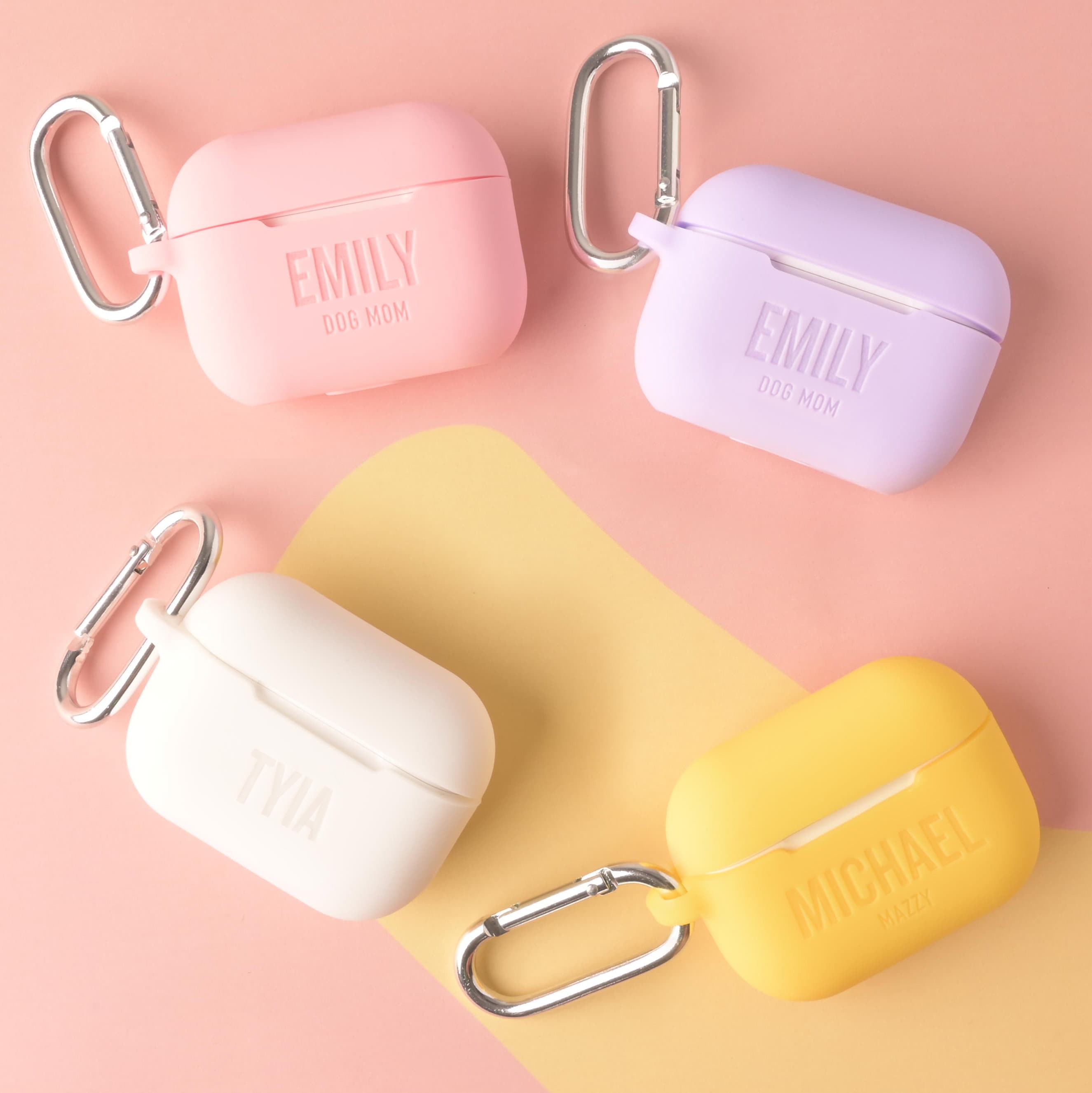 A collection of four personalized silicone AirPods Pro cases: pink and lavender cases engraved with 'EMILY DOG MOM,' a white case engraved with 'TYIA,' and a yellow case engraved with 'MICHAEL MAZZY.' All cases feature silver carabiner clips.