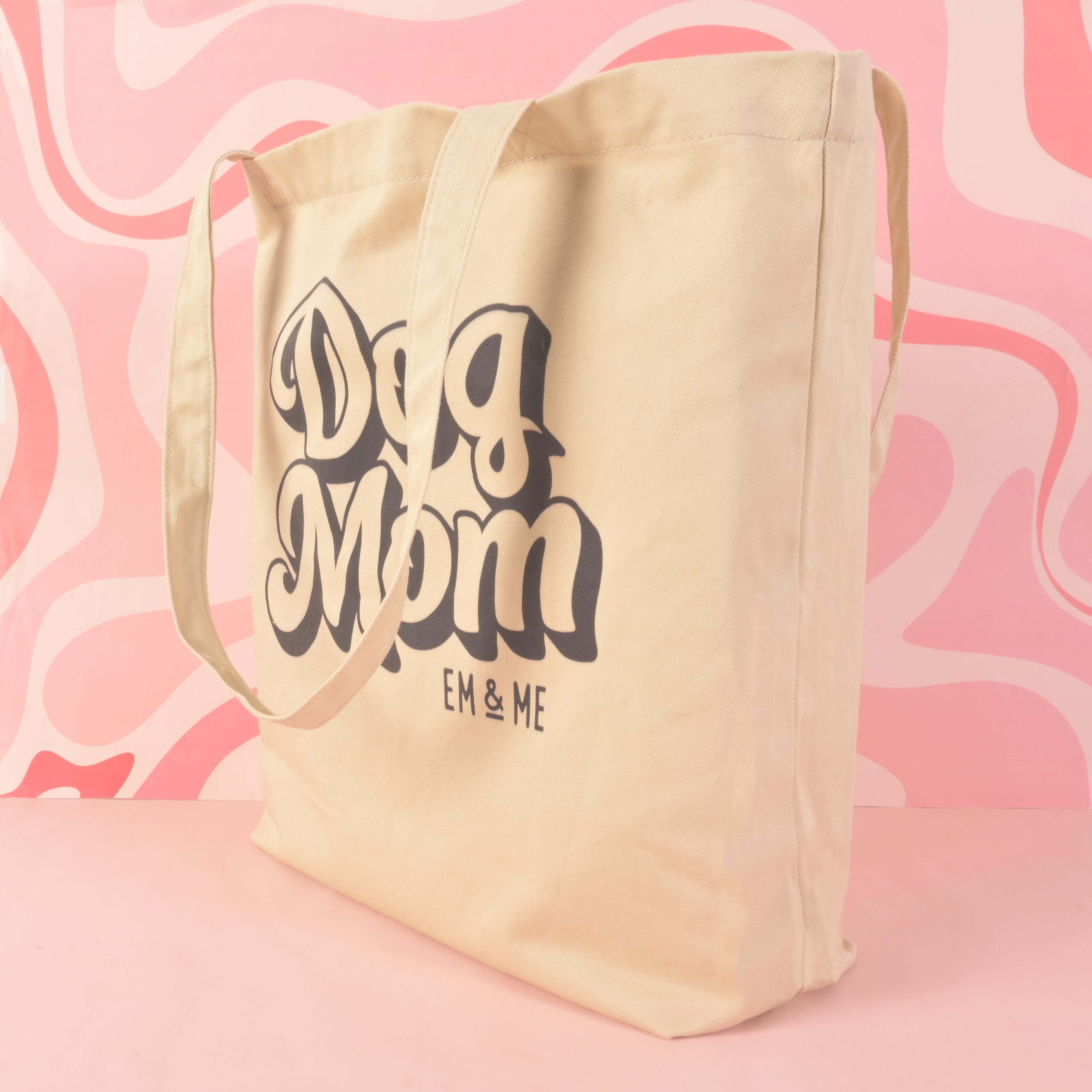 A product photo of a light-colored canvas tote bag shown from a side angle. The front of the bag is printed with a retro-style graphic that reads "Dog Mom" and "EM & ME." The bag is positioned on a pink background with a swirling, wavy pattern.